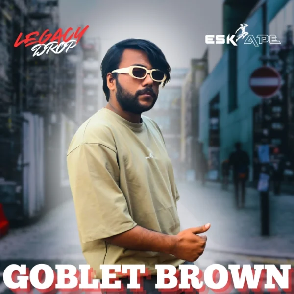 A man wearing a beige oversized t-shirt labeled "Goblet Brown" from the "Legacy Drop" collection by Eskape, posing in an urban setting while wearing white-framed sunglasses. Caption: The Goblet Brown t-shirt from the Legacy Drop collection by Eskape. Stylish, comfortable, and available now!