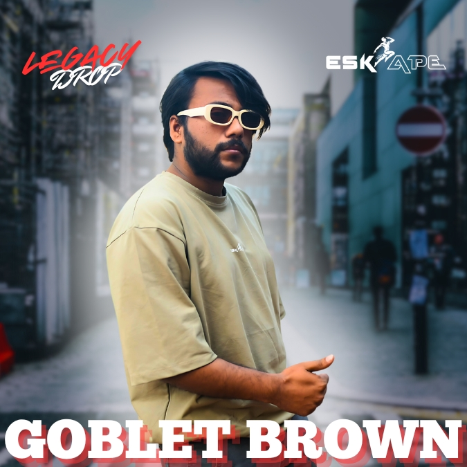 A man wearing a beige oversized t-shirt labeled "Goblet Brown" from the "Legacy Drop" collection by Eskape, posing in an urban setting while wearing white-framed sunglasses. Caption: The Goblet Brown t-shirt from the Legacy Drop collection by Eskape. Stylish, comfortable, and available now!