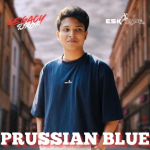 A young man wearing a deep blue oversized t-shirt labeled "Prussian Blue" from the "Legacy Drop" collection by Eskape, standing confidently in a stylish cityscape.