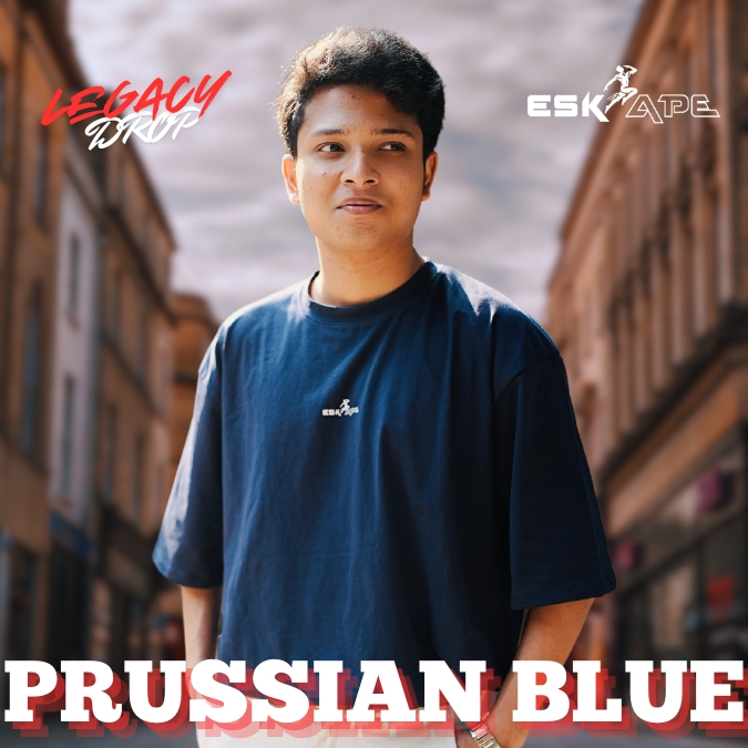 A young man wearing a deep blue oversized t-shirt labeled "Prussian Blue" from the "Legacy Drop" collection by Eskape, standing confidently in a stylish cityscape.