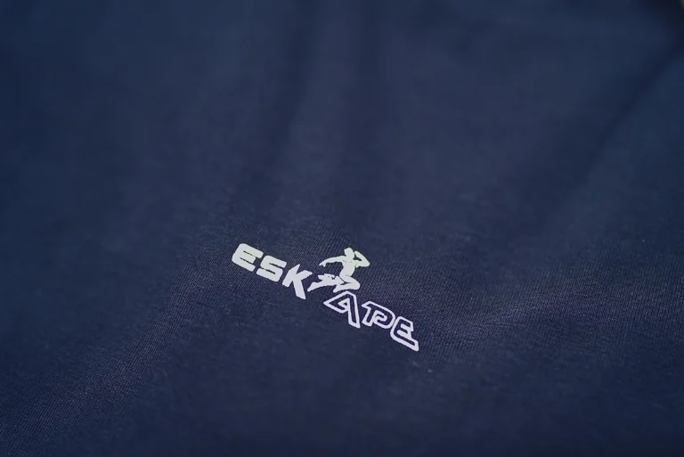 A close-up shot of the ESKAPE logo embroidery on a navy blue oversized drop-shoulder t-shirt, highlighting the premium fabric texture and minimalistic branding. The sleek and modern logo represents ESKAPE’s luxury streetwear identity.