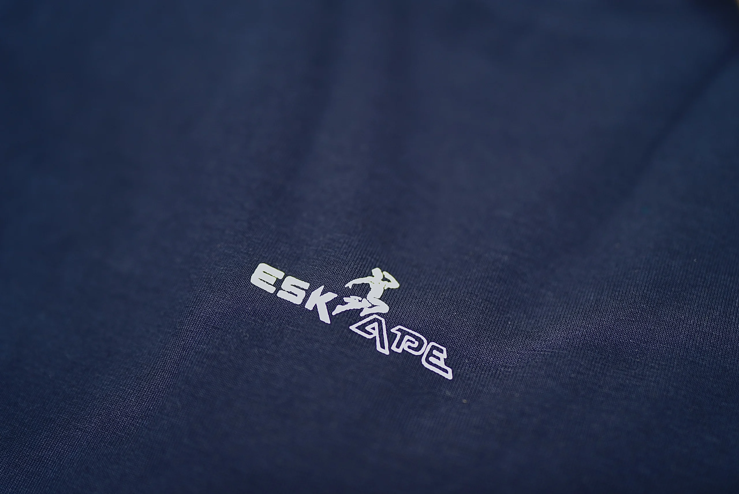 A close-up shot of the ESKAPE logo embroidery on a navy blue oversized drop-shoulder t-shirt, highlighting the premium fabric texture and minimalistic branding. The sleek and modern logo represents ESKAPE’s luxury streetwear identity.