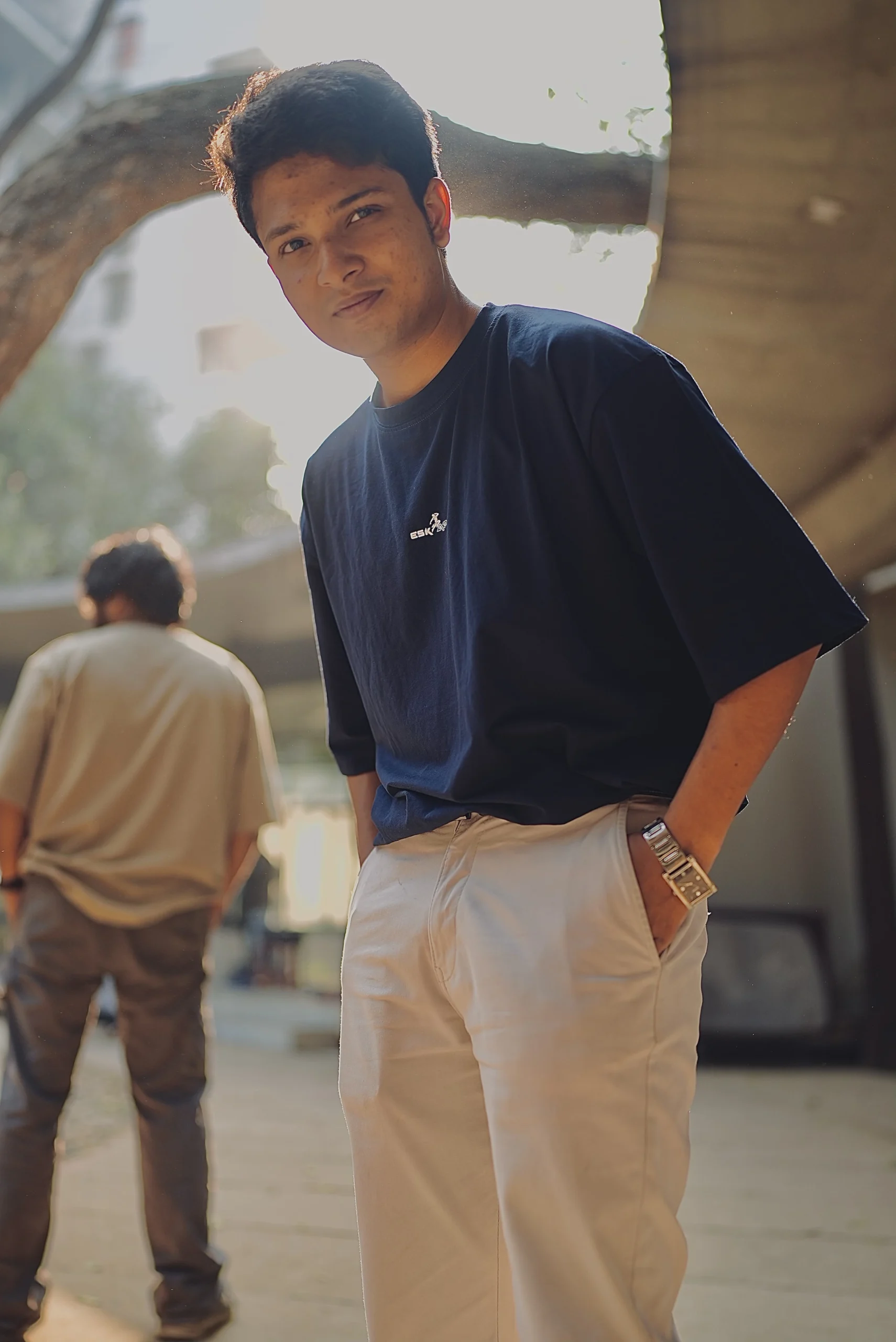 A young man confidently poses in a navy blue ESKAPE oversized drop-shoulder t-shirt paired with beige trousers, exuding a clean and minimal luxury streetwear vibe. Another individual in a beige ESKAPE t-shirt is seen in the background, adding depth to the urban, fashion-forward aesthetic.