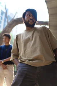 A young man in a beige ESKAPE oversized drop-shoulder t-shirt poses confidently from a low-angle perspective, exuding a bold and fearless attitude. Another individual in a navy-blue ESKAPE t-shirt stands in the background, adding depth to the urban, streetwear aesthetic.