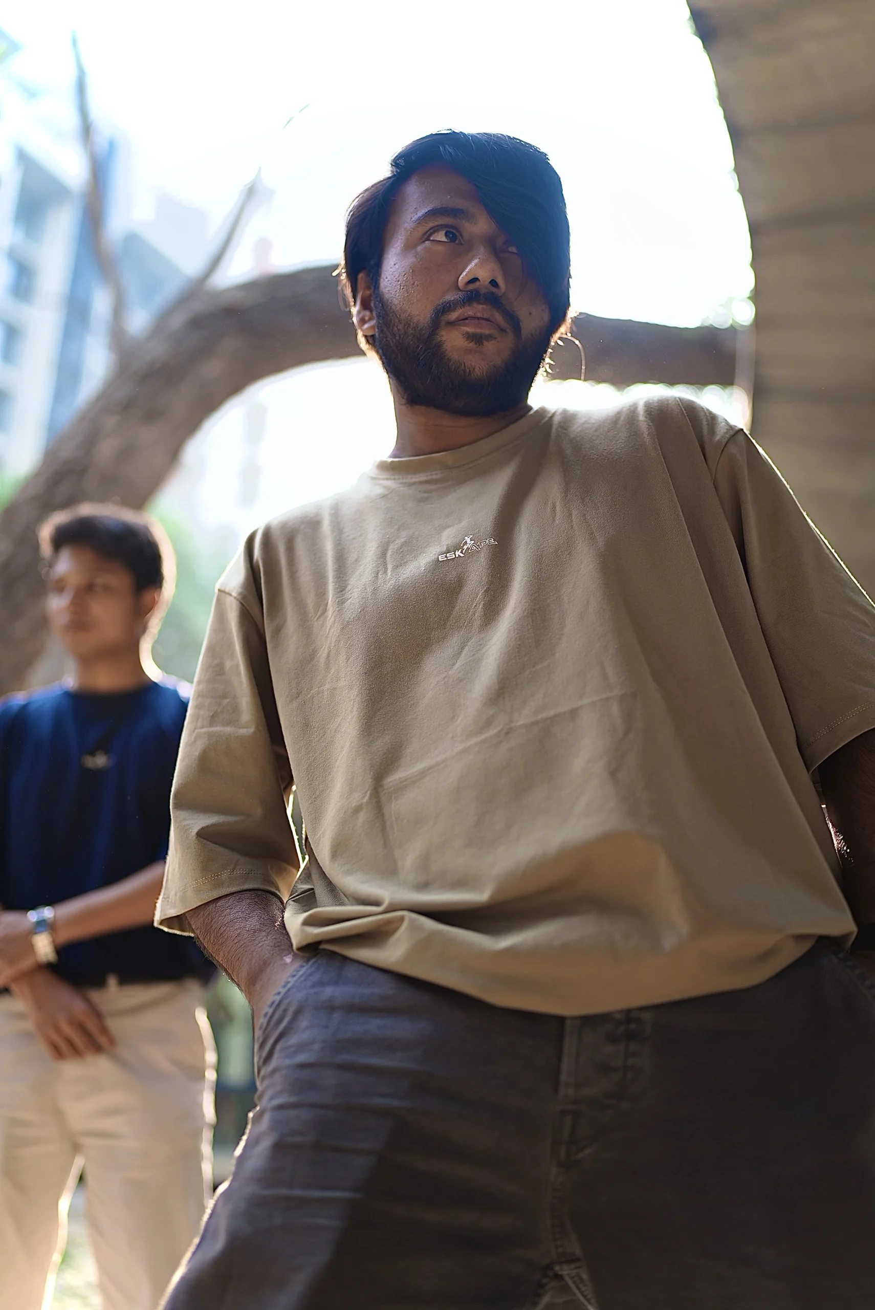 A young man in a beige ESKAPE oversized drop-shoulder t-shirt poses confidently from a low-angle perspective, exuding a bold and fearless attitude. Another individual in a navy-blue ESKAPE t-shirt stands in the background, adding depth to the urban, streetwear aesthetic.