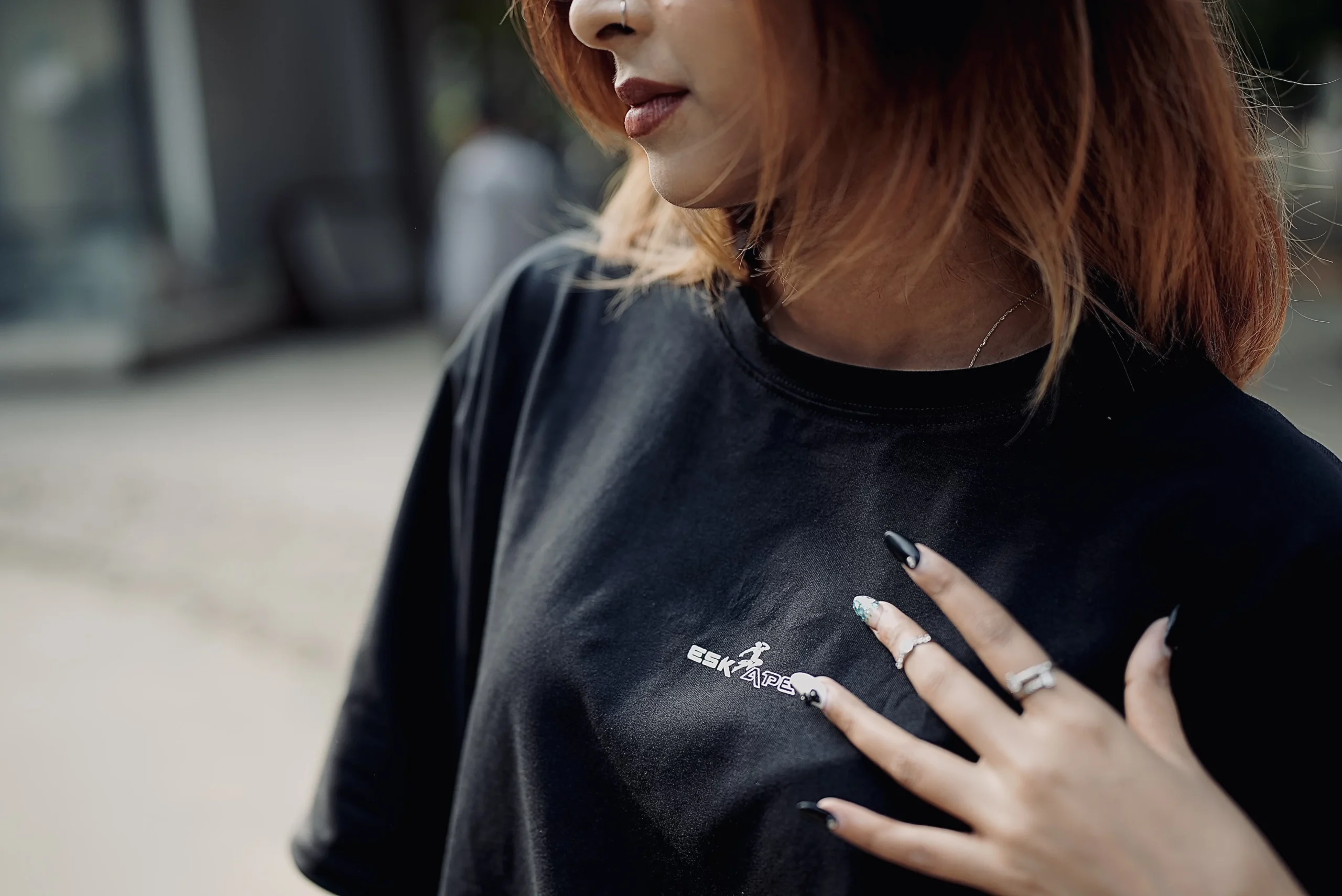 A close-up shot of a young woman wearing a black ESKAPE oversized drop-shoulder t-shirt, highlighting the ESKAPE logo embroidery on the chest. Her well-manicured nails and accessories add to the luxury streetwear aesthetic, making a bold fashion statement.