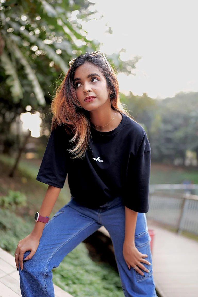 A stylish young woman wearing a black ESKAPE oversized drop-shoulder t-shirt and blue cargo jeans poses confidently in an outdoor setting with lush greenery in the background. Her bold, effortless fashion style embodies ESKAPE’s premium streetwear aesthetic.