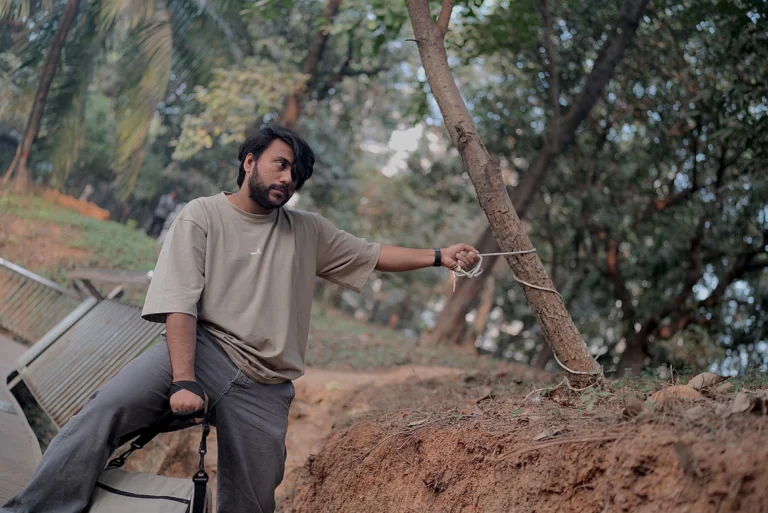 A stylish young man wearing a beige ESKAPE oversized drop-shoulder t-shirt and relaxed-fit pants sits outdoors in a rugged, natural setting. He holds a rope tied to a leaning tree, capturing the essence of ESKAPE’s adventurous, bold, and free-spirited fashion identity.