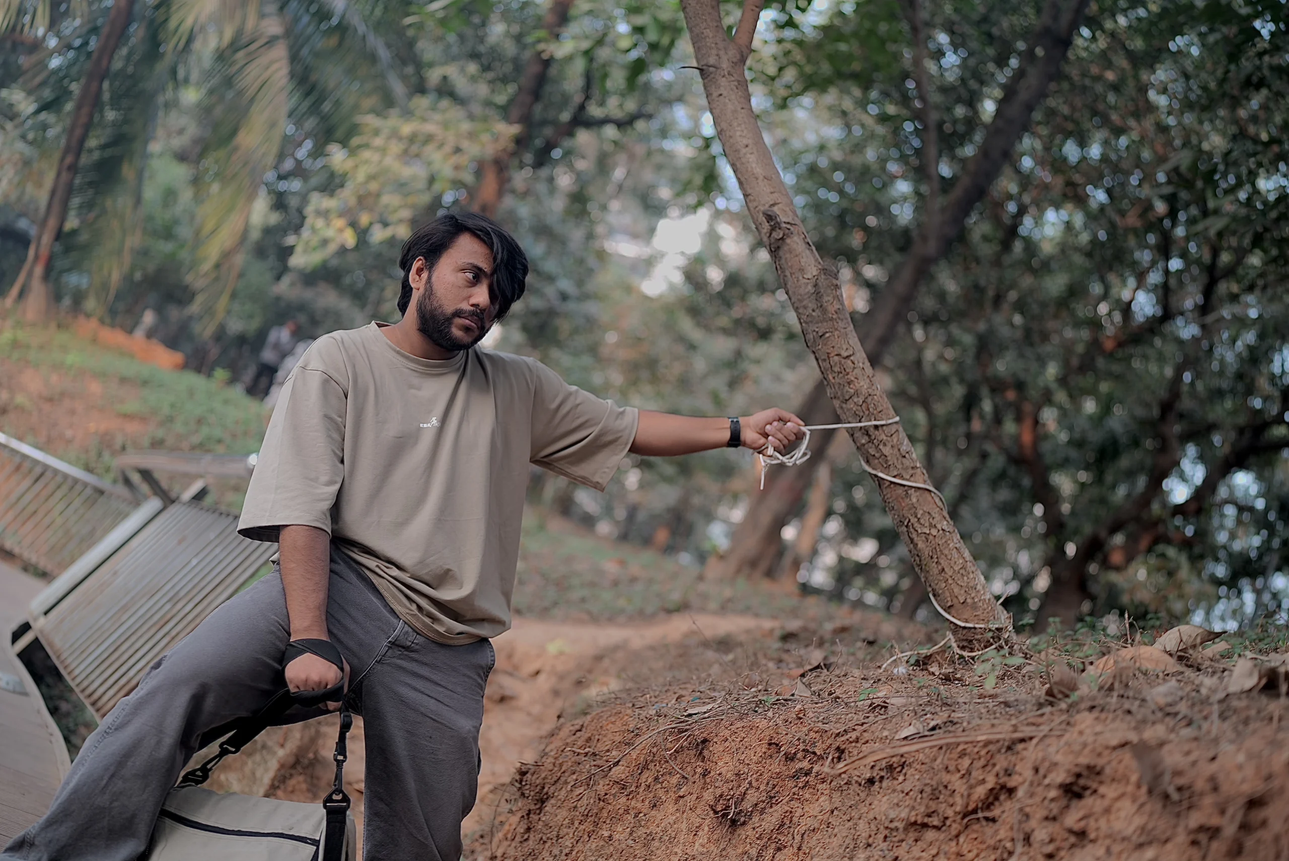 A stylish young man wearing a beige ESKAPE oversized drop-shoulder t-shirt and relaxed-fit pants sits outdoors in a rugged, natural setting. He holds a rope tied to a leaning tree, capturing the essence of ESKAPE’s adventurous, bold, and free-spirited fashion identity.