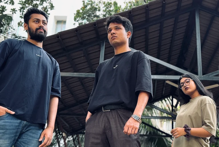 A group of three individuals wearing ESKAPE oversized drop-shoulder t-shirts in black and beige stand confidently in an urban setting with industrial architecture in the background. Their bold expressions and modern streetwear style reflect ESKAPE’s premium and fearless fashion identity.