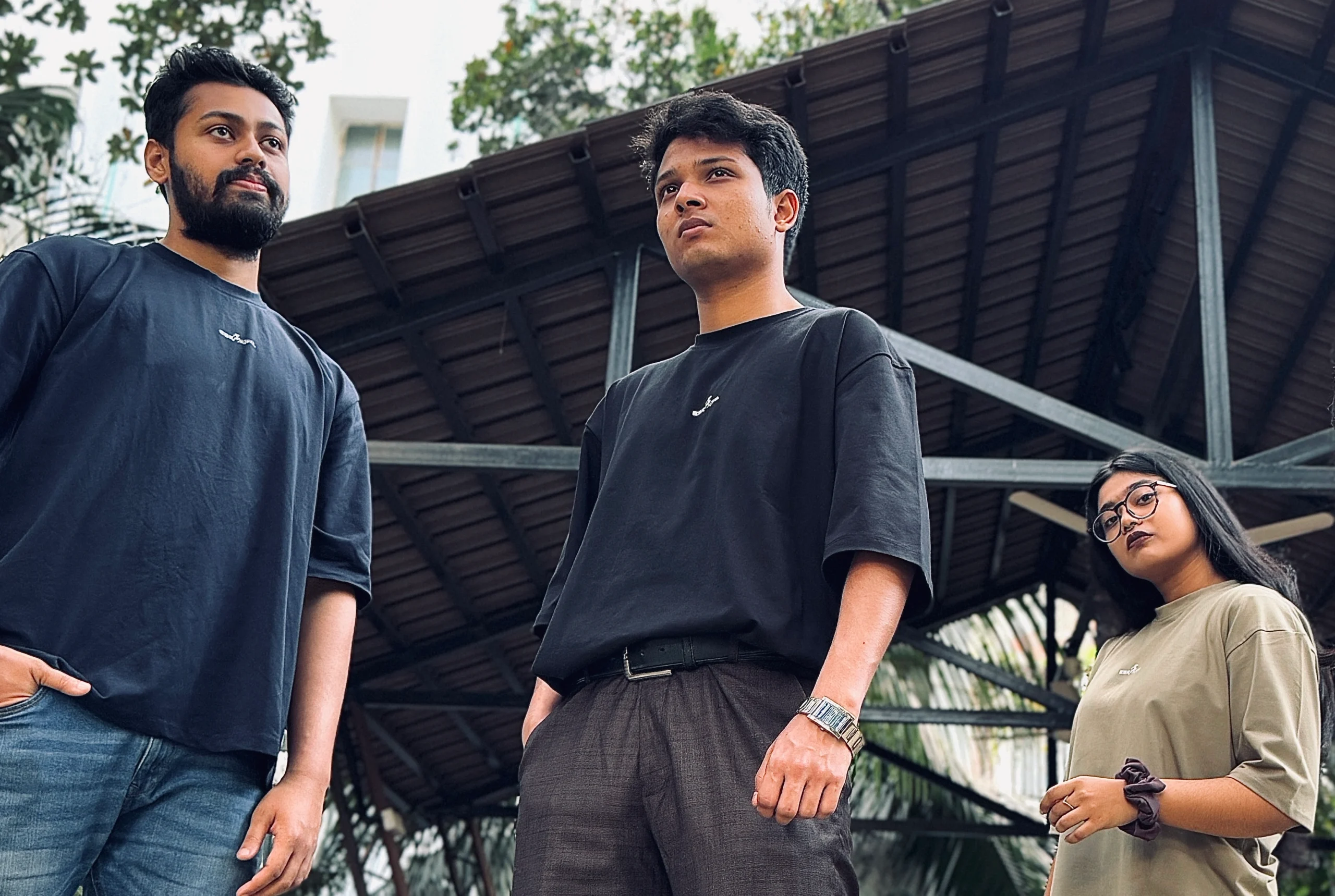 A group of three individuals wearing ESKAPE oversized drop-shoulder t-shirts in black and beige stand confidently in an urban setting with industrial architecture in the background. Their bold expressions and modern streetwear style reflect ESKAPE’s premium and fearless fashion identity.