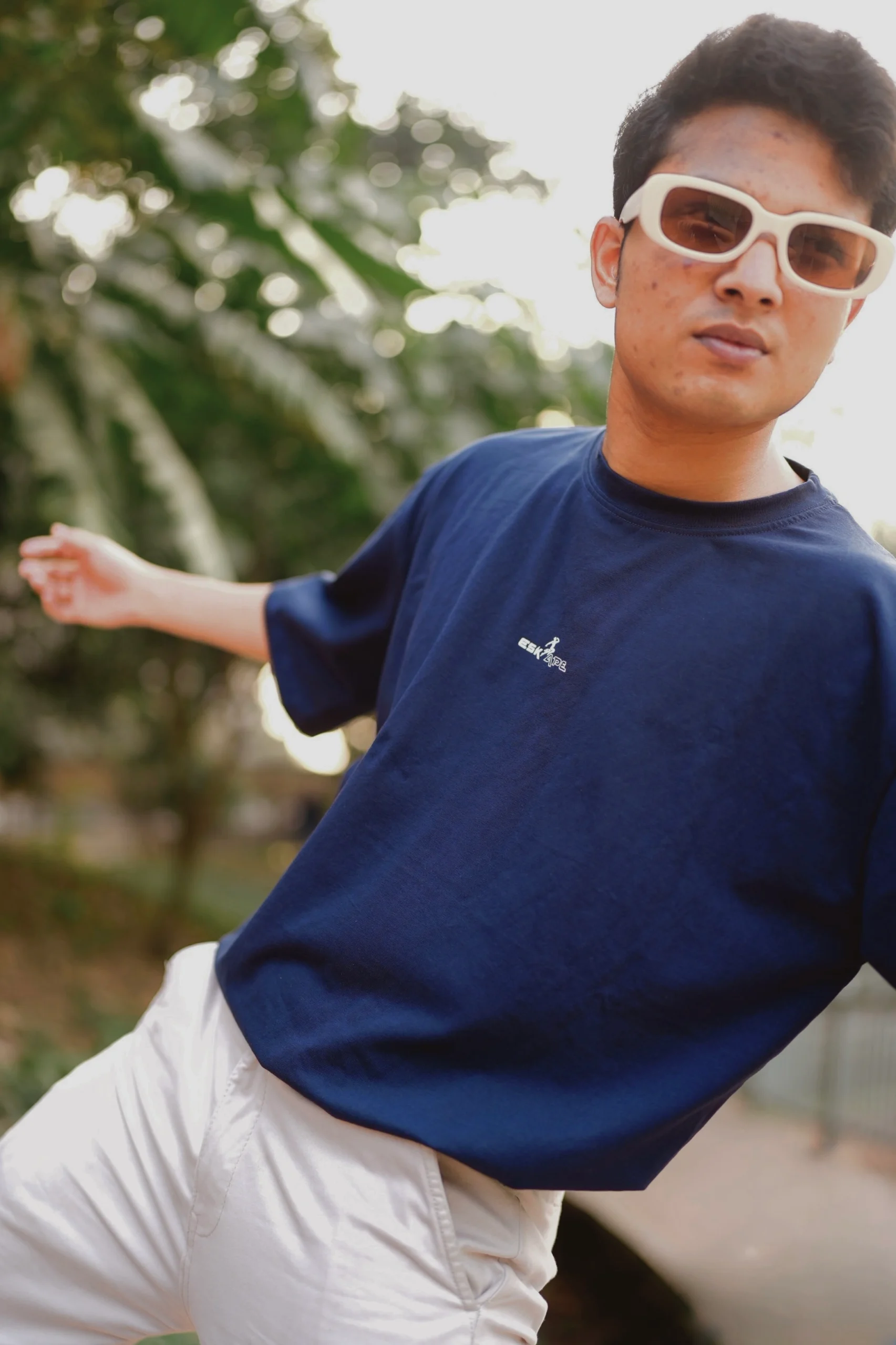 A young man wearing a navy blue ESKAPE oversized drop-shoulder t-shirt and white trousers strikes a dynamic pose outdoors. His bold white sunglasses and confident expression embody ESKAPE’s fearless, statement-making streetwear aesthetic.A young man wearing a navy blue ESKAPE oversized drop-shoulder t-shirt and white trousers strikes a dynamic pose outdoors. His bold white sunglasses and confident expression embody ESKAPE’s fearless, statement-making streetwear aesthetic.