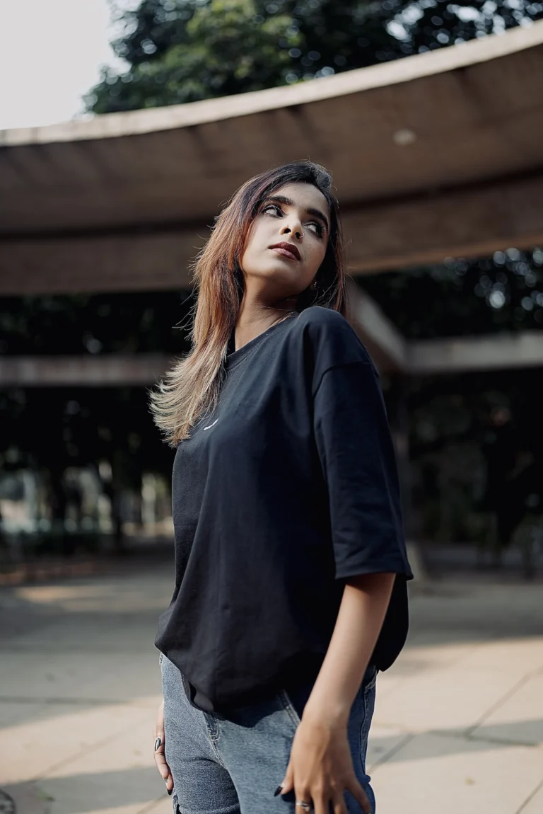 A young woman confidently poses in a black ESKAPE oversized drop-shoulder t-shirt paired with blue cargo jeans. The urban backdrop and her poised expression embody ESKAPE’s bold, minimal, and premium streetwear identity.
